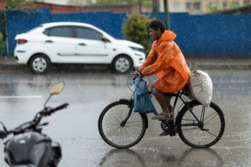 Radfahrer mit orangefarbener Regenjacke fährt im Starkregen neben einem Auto
